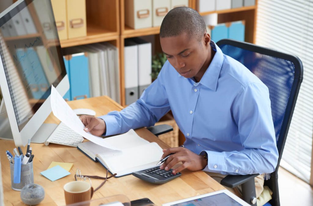 homem-negro-usand-camisa-azul-claro-realizando-tributacao-para-ecommerce-em-escritorio-com-livros-folhas-e-computador