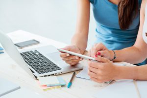 Imagem de duas mulheres sentadas em uma mesa branca, ao lado de um celular, um notebook e vários post-it e canetas, lendo o post de “Cadastro de produtos na loja online: veja como deve ser feito” para aprenderem como cadastrar seus produtos em sua loja virtual.