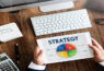 maos-segurando-grafico-de-estrategias-de-marketing-para-loja-virtual-sobre-mesa-de-escritorio-com-teclado-de-computador