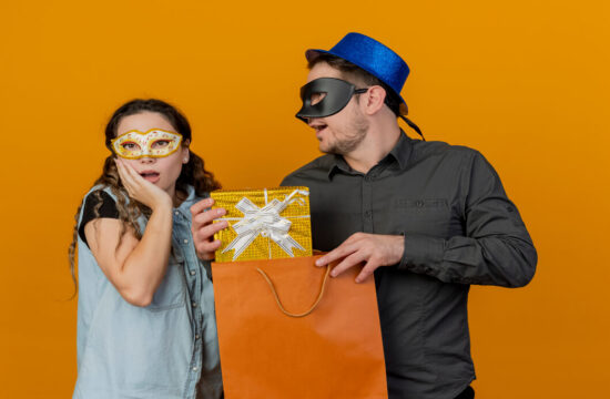 homem-e-mulher-trocam-sacolas-de-compras-com-mascara-de-carnaval-sobre-fundo-laranja