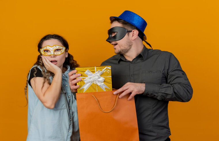 homem-e-mulher-trocam-sacolas-de-compras-com-mascara-de-carnaval-sobre-fundo-laranja