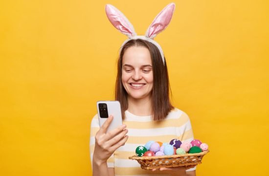 mulher-alegre-usando-orelhas-de-coelho-segurando-ovos-de-pascoa-ecommerce-sobre-fundo-amarelo-com-celular