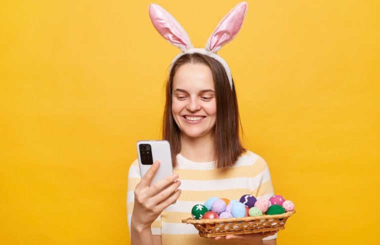 mulher-alegre-usando-orelhas-de-coelho-segurando-ovos-de-pascoa-ecommerce-sobre-fundo-amarelo-com-celular