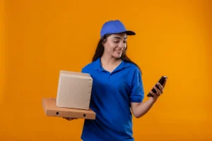jovem-entregadora-de-uniforme-azul-e-bone-segurando-caixas-de-papelao-olhando-para-a-tela-do-smartphone-sorrindo-em-pe-sobre-fundo-amarelo