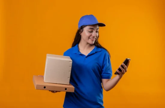 jovem-entregadora-de-uniforme-azul-e-bone-segurando-caixas-de-papelao-olhando-para-a-tela-do-smartphone-sorrindo-em-pe-sobre-fundo-amarelo